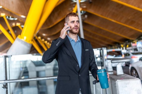 Businessman in a suit holding a reusable coffee cup while using a smartphone, waiting at the