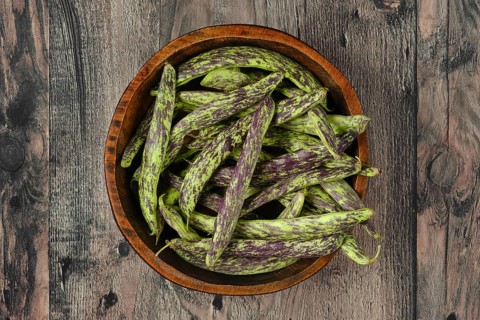 A wooden bowl filled to the brim with vibrant purple and green beans rests on a textured wooden