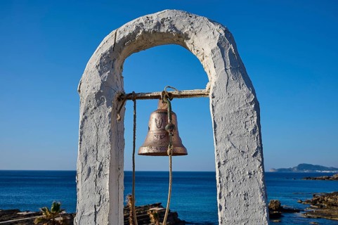 Close-up of a bell in a white frame in front of a rocky coastal landscape and blue sky, rock church