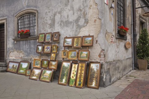 Framed paintings displayed against an old damaged grey cement wall in the Old Town Market Square,