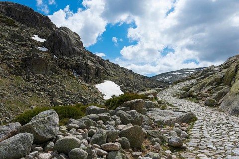 A stony path through a rocky mountain region with patches of snow under a cloudy sky, hiking area,