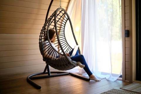 On a hot sunny day, a young woman sits in a hanging swing chair with a laptop in an air-conditioned