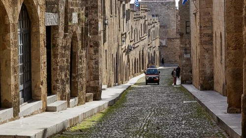 A historic cobblestone street with old yellow stone buildings, a moving car and a pedestrian,
