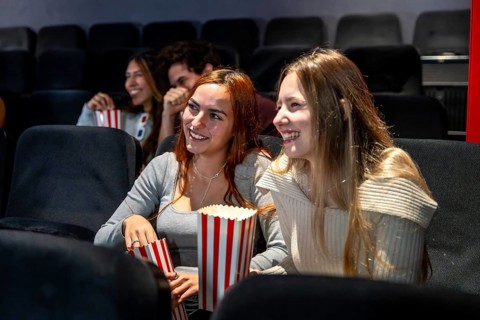 Two young women enjoying popcorn and smiling while watching a movie together in a cozy cinema,
