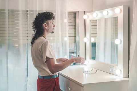 Young man with a mullet hairstyle standing in front of a lit mirror, holding items, dressed in a