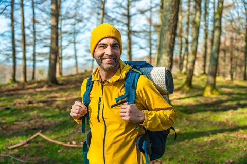 Hiker in a bright yellow jacket and backpack smiling while trekking through a sunlit forest,