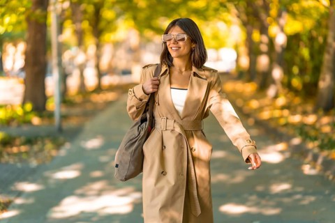 A woman wearing a tan trench coat and sunglasses walks down a sidewalk. She is carrying a brown
