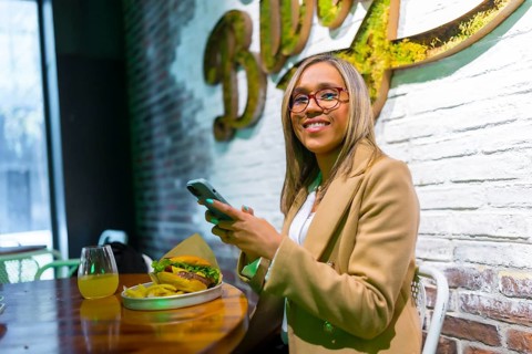 Latin woman smiling at camera sitting on a restaurant of fast food eating cheeseburger while using