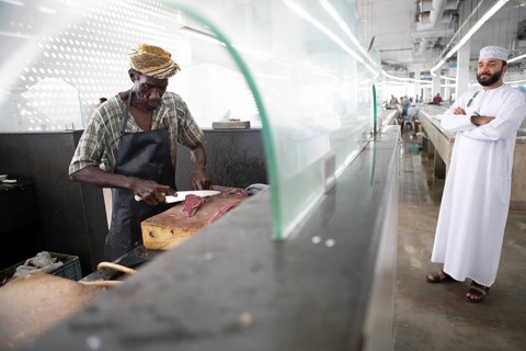 Omani man filleting a fish in Muttrah fish market, an Omani man in dishdasha watches, Muscat, Oman,