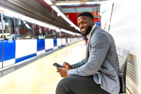 Young black businessman sitting on bench in subway station using smartphone while waiting for train