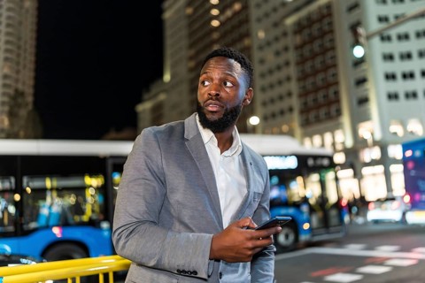 Young businessman holding a smartphone, looking around in a bustling city at night, surrounded by