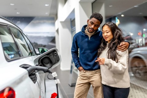Multiethnic couple examining charging port of electric vehicle at car dealership, embracing