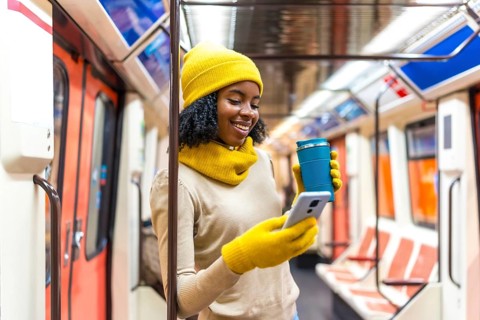 Smiling woman wearing yellow winter clothes using phone and holding reusable coffee cup while