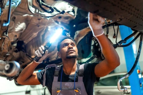 Young mechanic using flashlight, inspecting underside of vehicle raised on lift in professional