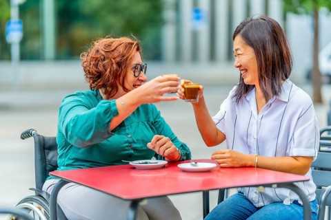 Woman with disability and asian friend toasting with coffee sitting on an outdoor cafeteria in the