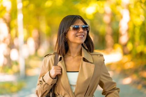 A woman wearing sunglasses and a brown coat is walking down a path. She is smiling and holding a