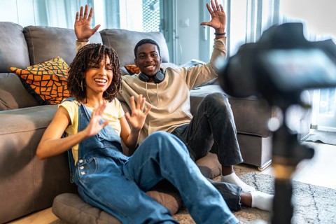 Two influencers happily waving at the camera while recording a video blog from their cozy living