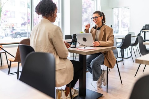 Two women engaging in a podcast recording session at a cafe, utilizing a laptop and microphone for