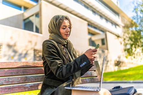 Muslim businesswoman using mobile working on remote with a laptop sitting on a bench in a park