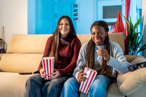 Two happy teenage girls eating popcorn while watching a movie at home, sharing a moment of leisure