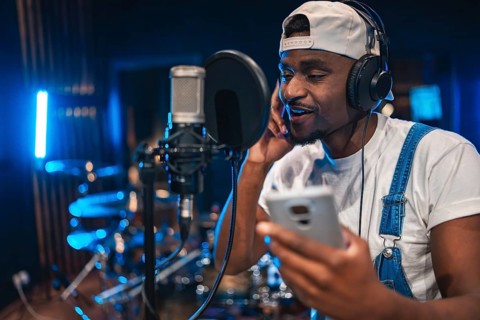 Young man recording vocals in professional music studio using smartphone, microphone and headphones