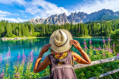 Woman with backpack and straw hat admiring the turquoise waters of lake carezza and the latemar