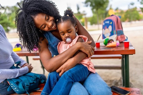 Happy african woman embracing her baby daughter in the park sitting on a picnic table