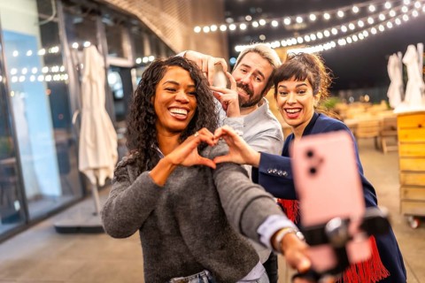 Multi-ethnic friends join hands gesturing heart shape while vlogging online in a night party