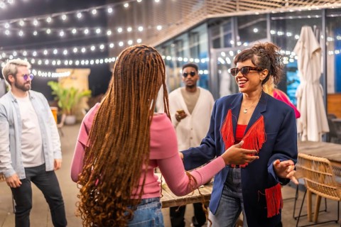 Two multi-ethnic friends women dancing together next to other friends at night party outdoors