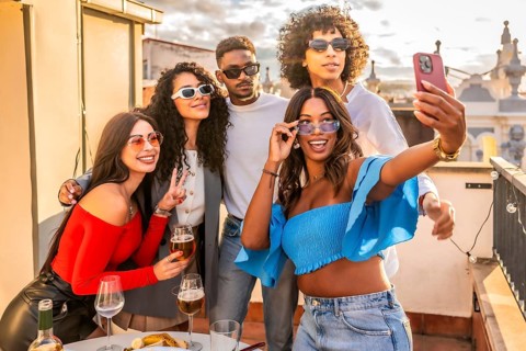 Woman taking selfie with multi-ethnic friends in a roof party with wine and snacks during sunset