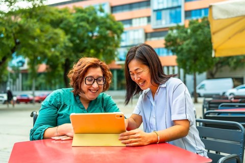 Disabled woman in electric wheelchair and chinese female friend using tablet sitting on sidewalk
