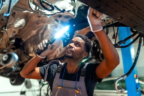 Young mechanic using flashlight, inspecting underside of vehicle raised on lift in car workshop