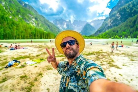 Tourist wearing sunglasses and straw hat gesturing peace sign while taking a selfie at the beach of