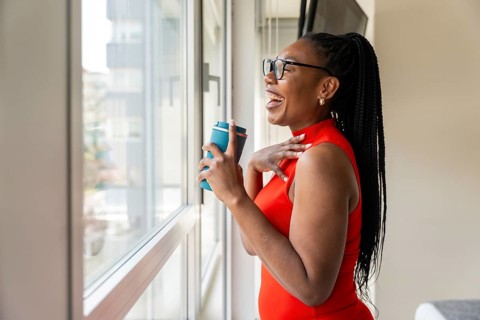 Happy businesswoman laughing while holding a reusable coffee cup and looking out the window of her