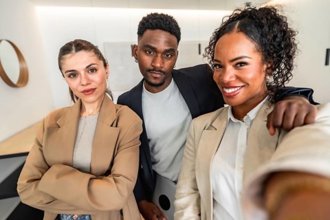 Three smiling businesspeople are taking a selfie in a modern office, showing teamwork and