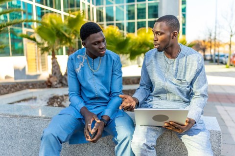 Two senegalese businessmen wearing traditional dashiki attire are sitting outside a modern office
