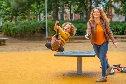 Caucasian adult mother and girl having fun swinging in a playground in an urban park