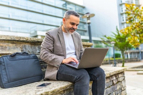Happy mature caucasian businessman using laptop sitting on a stone bench outdoors in the financial