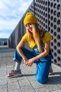 Blonde dancer wearing yellow beanie, yellow t shirt and blue dungarees, kneeling and posing in a