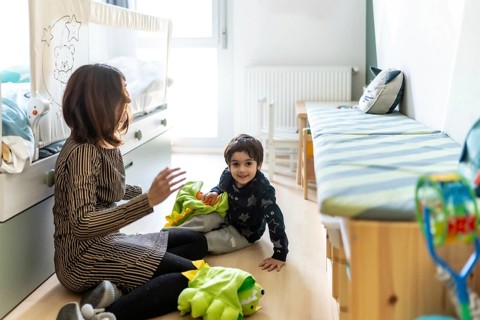 Mother and son are sitting on the floor, playing with a dinosaur costume in a brightly lit bedroom