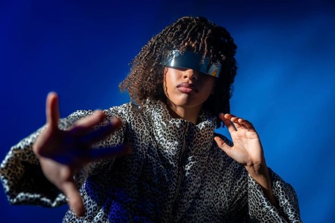 Stylish person with futuristic visor and patterned jacket extending hand on vibrant blue background
