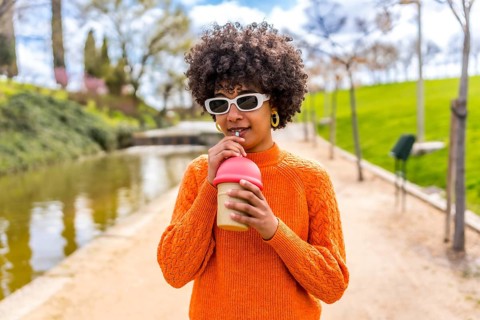 Stylish young woman with an afro hairstyle sips a beverage through a straw in a serene park setting