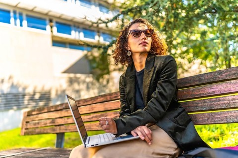 Modern arab businesswoman using laptop sitting on park bench working on remote