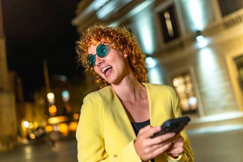 Modern woman in colorful casual clothes and blue sunglasses using smartphone laughing in the city