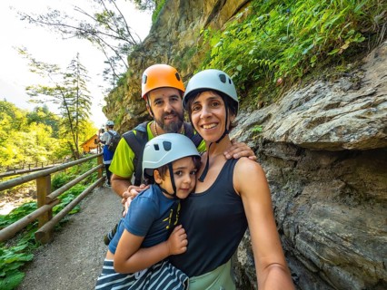 Family wearing helmets taking selfie while hiking on wooden walkway next to rock wall in beautiful