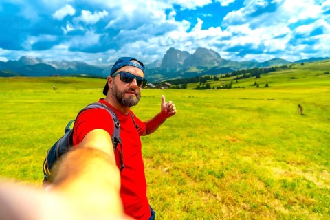 Backpacker taking selfie showing thumbs up sign enjoying beautiful summer landscape at alpe di
