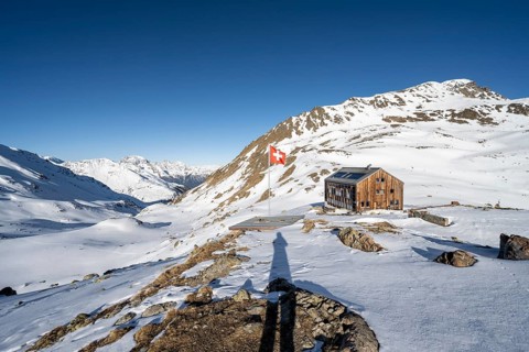 Swiss flag and mountain hut Keschhütte in front of mountain landscape in winter, behind summit of