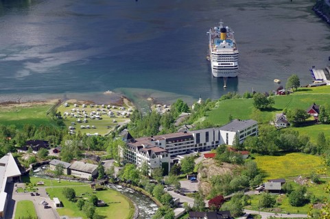 A large cruise ship is moored on a shore with many buildings and green meadows and trees, Geiranger