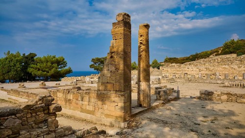 Two ancient stone columns stand in the middle of a ruined site under a slightly cloudy sky,