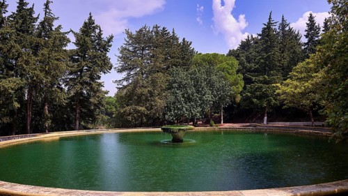 A round lake with a central fountain surrounded by tall green trees under a blue sky with clouds,